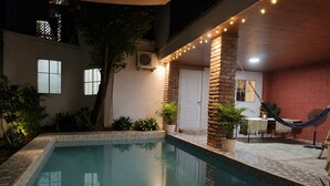 Una piscina con borde de azulejos, rodeada de plantas en macetas y una hamaca.
