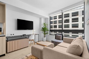 Un salon moderne avec un canapé, une table basse, un téléviseur fixé au mur et une fenêtre donnant sur un immeuble.