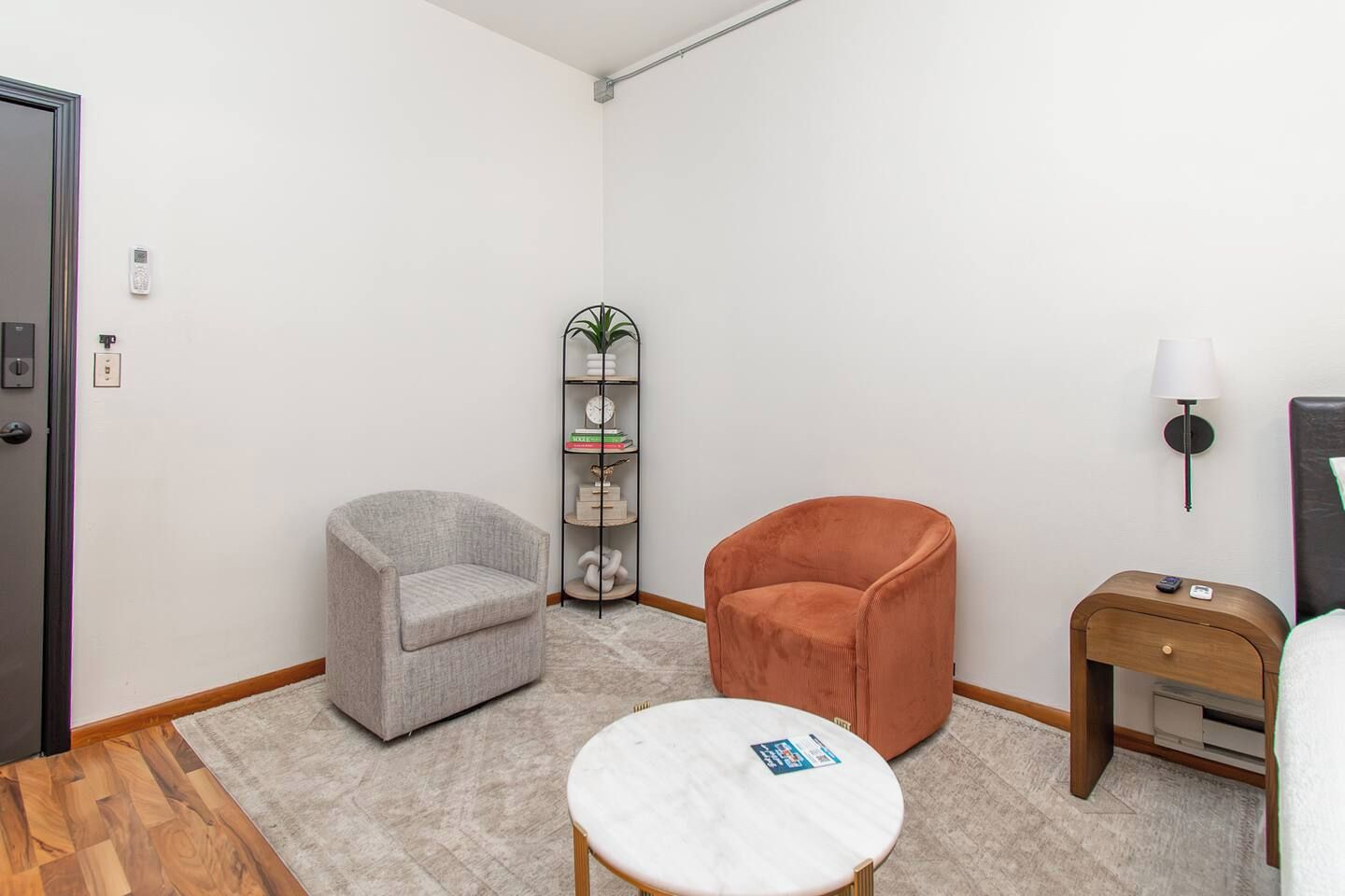 Een hotelkamer met een grijze fauteuil, een oranje fauteuil, een klein rond tafeltje, een boekenkast en een nachtkastje met een lamp.