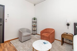 Een hotelkamer met een grijze fauteuil, een oranje fauteuil, een klein rond tafeltje, een boekenkast en een nachtkastje met een lamp.