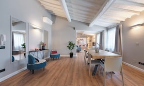 Amplia sala de estar con piso de madera, una mesa de comedor y un conjunto de asientos. Se observa un espejo grande a la izquierda y una planta en el centro.