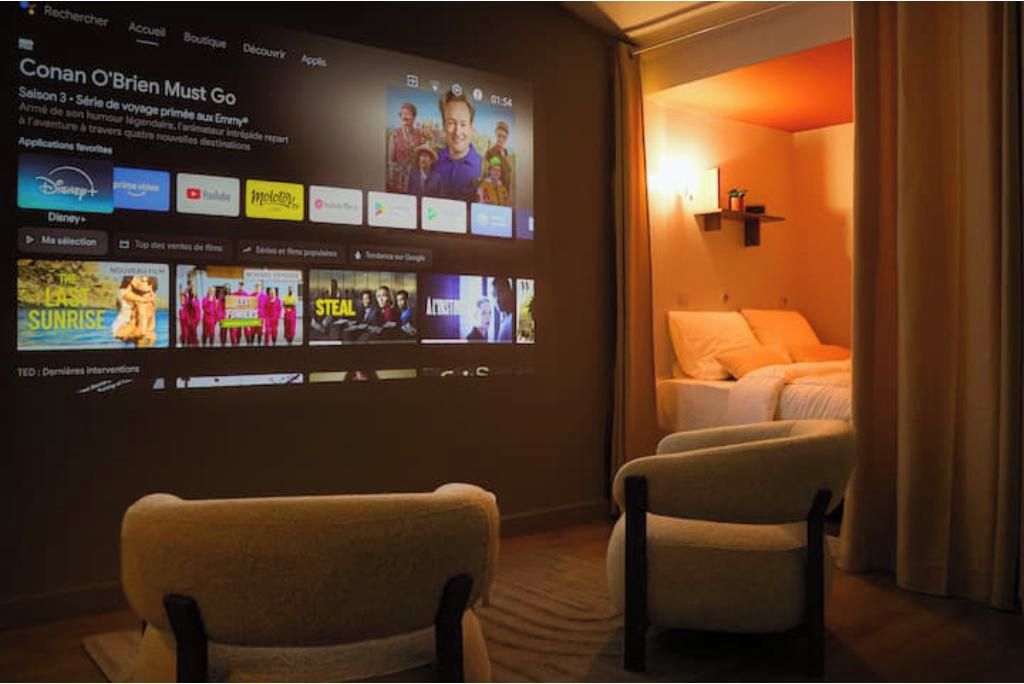 A hotel room with a large screen displaying a movie, two armchairs, a bed, and a wall-mounted shelf.