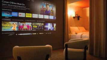 A hotel room with a large screen displaying a movie, two armchairs, a bed, and a wall-mounted shelf.