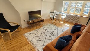 Ett vardagsrum i loftstil med trägolv, en soffa, en tv, ett matbord med stolar och ett stort fönster.