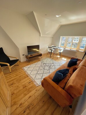 Un salón estilo loft con piso de madera, un sofá, un televisor, una mesa de comedor con sillas y un ventanal grande.