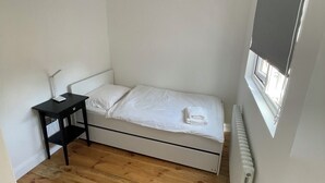 A small, single-bed room with a wooden floor, a bedside table, and a window with a blind. - Nice London flat with free private parking and 20 min train to central London (Bromley)