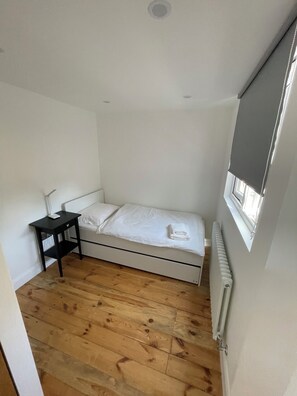 Ein kleines Zimmer mit einem Einzelbett, Holzboden, einem Nachttisch und einem Fenster mit Jalousie.