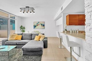 Ein modernes Wohnzimmer mit einem grauen Ecksofa, einem Glastisch und einem Essbereich mit weißen Barhocker.