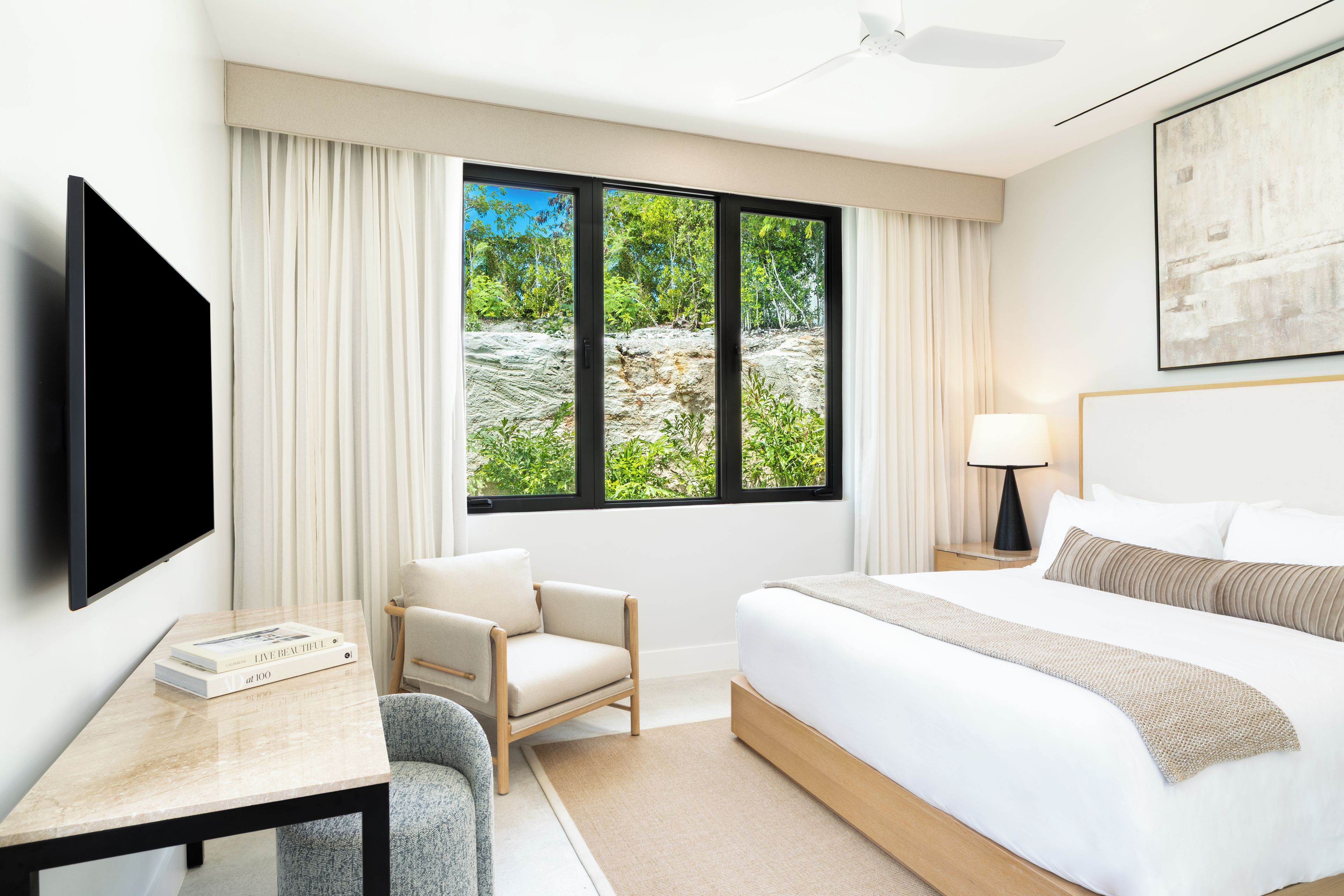 A hotel room with a bed, a chair, a desk, a TV, and a window with a view of greenery.