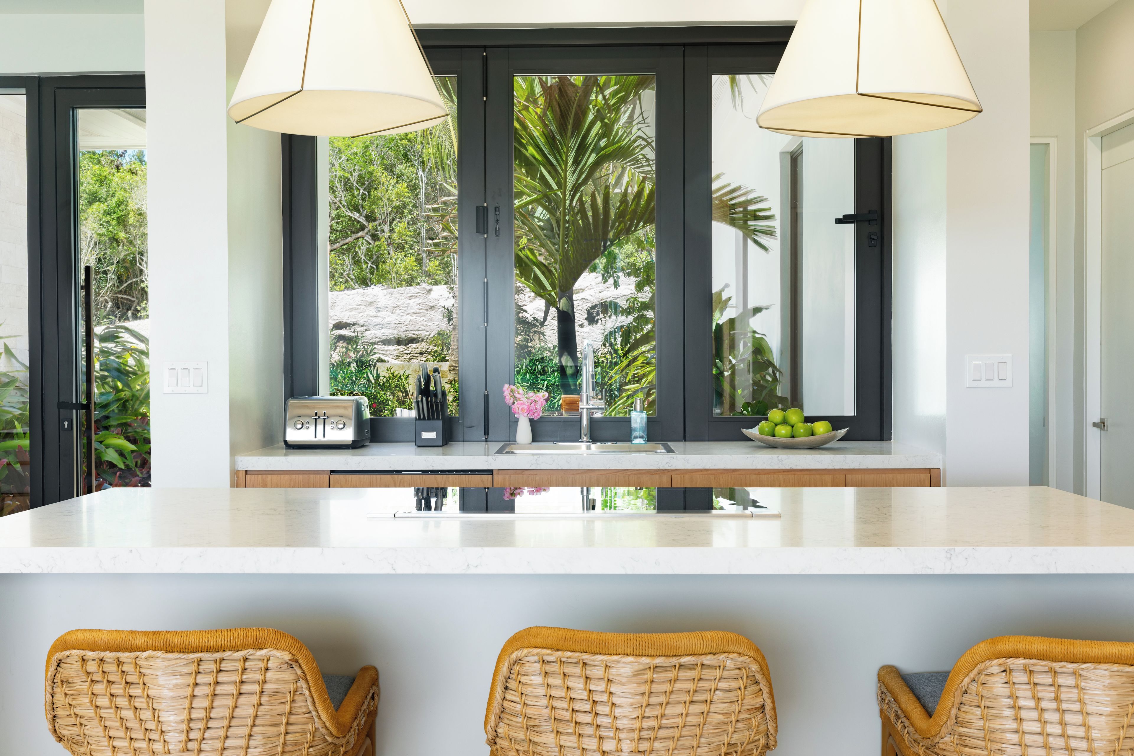 A modern kitchen with a large island, two pendant lights, and a view of greenery outside.