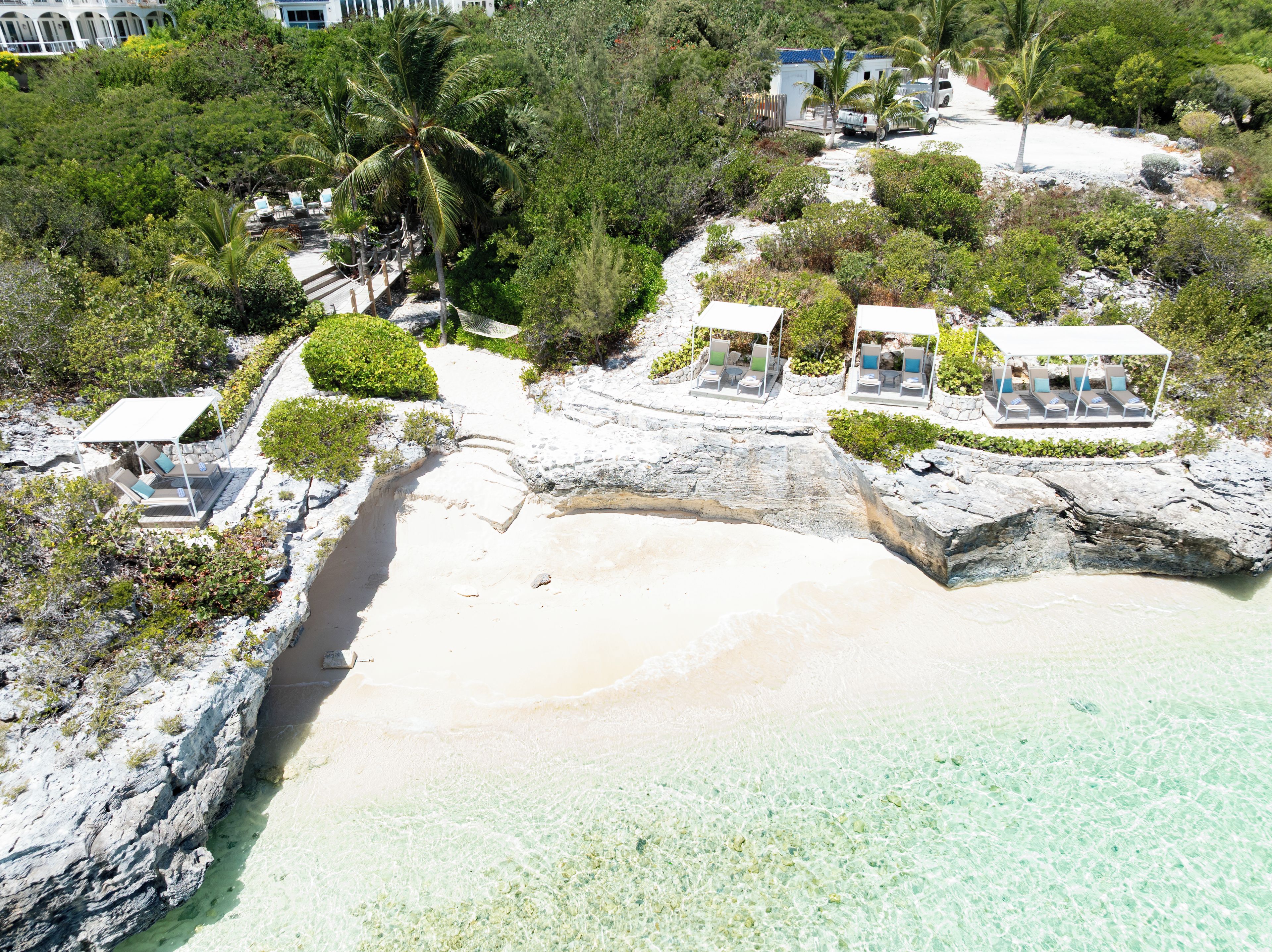 A coastal property with a rocky beach, clear waters, and a series of outdoor seating areas with umbrellas.