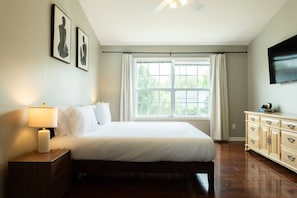 Une chambre à coucher comprenant un lit, une table de chevet, une commode, une fenêtre avec des rideaux et un téléviseur à écran plat fixé au mur.