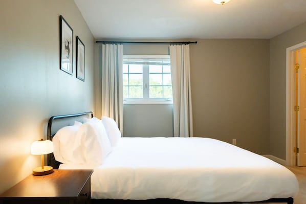 Una habitación de hotel con cama, mesita de noche con lámpara, ventana con cortinas y puerta.