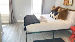 Un dormitorio con cama, almohadas, una colcha, una silla roja y una ventana con cortinas.