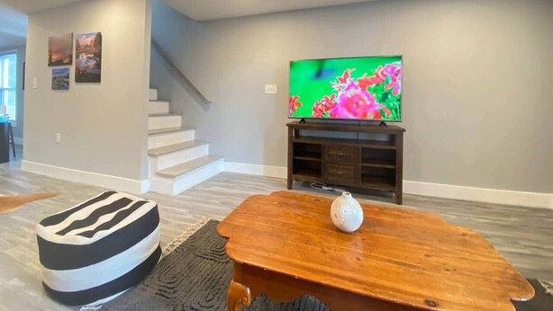 Una sala de estar con una televisión de pantalla plana sobre un mueble bajo, una mesa de centro de madera y un reposapiés a rayas.