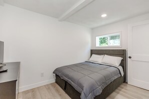 A bedroom with a bed, a window, a door, and a desk with a remote control.