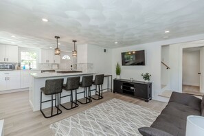 A modern kitchen with white cabinets, a central island with bar stools, pendant lighting, and a flat-screen TV mounted on the wall.