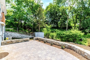 Un espace extérieur pavé, agrémenté d’un muret en pierre, et entouré d’une végétation luxuriante et d’arbres.