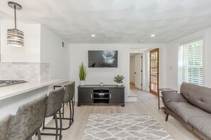 A modern living room with a grey sofa, a dining area with a bar, and a TV mounted on the wall.