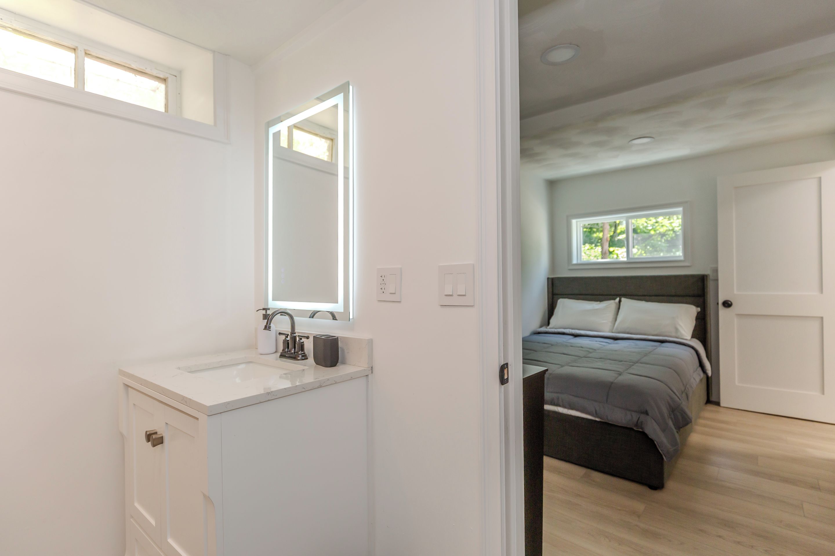 Quarto com cama, janela e porta que dá para um banheiro com pia e espelho.