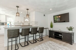A modern kitchen with a central island, bar stools, pendant lighting, a flat-screen TV, and a potted plant.