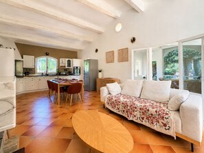 Eine moderne Küche mit weißen Schränken, einem Esstisch mit Stühlen, einer Couch mit Blumenkissen und einem großen Fenster mit Blick auf Grünflächen.
