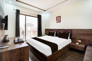 A hotel room with a large bed, a bedside table, a nightstand, a window with curtains, a wall-mounted TV, and a framed picture.