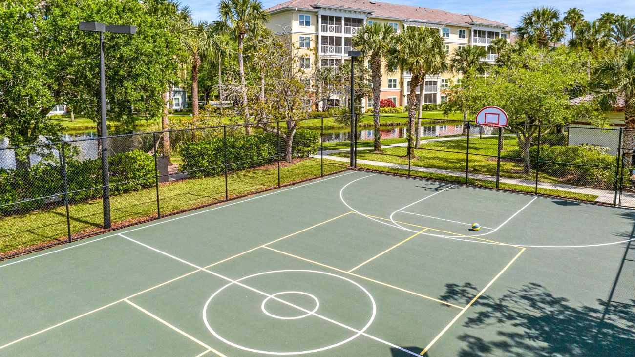 Una cancha de baloncesto al aire libre con una cerca, árboles y un edificio al fondo.