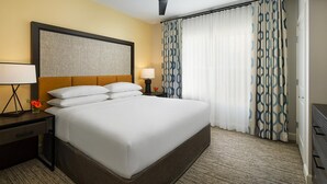 Ein Hotelzimmer mit einem großen Bett, einem Nachttisch mit Lampe, einem Wandbild und einem Fenster mit gemusterten Vorhängen.