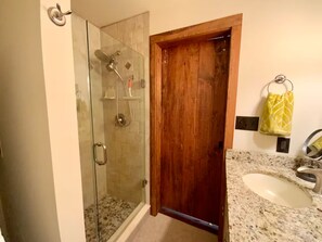 Une salle de bain avec une douche à parois de verre, une porte en bois, un lavabo avec un plan de travail en granit et une serviette jaune accrochée à un crochet.