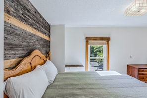Ein Schlafzimmer mit einem hölzernen Kopfteil, einem Bett mit weißer und grüner Bettwäsche, einer Holzkommode und einem Fenster mit Blick auf Grünflächen.