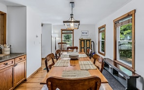 Een eetkamer met een lange houten tafel, stoelen, een kroonluchter en grote ramen.