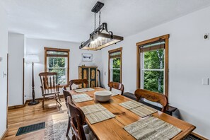 Ein Esszimmer mit Holztisch, Stühlen, einer Chandelüster und Fenstern mit Blick auf Bäume.