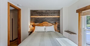 Een slaapkamer met een houten hoofdbord, een bed met witte beddengoed, een nachtkastje met een lamp en een raam met uitzicht op groen.