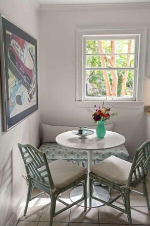 Quarto com parede branca, janela com vista para árvores, mesa redonda com um vaso de flores e duas cadeiras com almofadas.