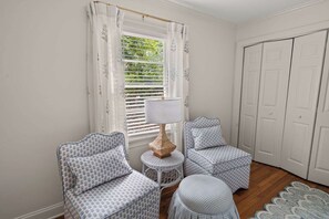 Quarto com janela com persianas, luminária, duas cadeiras estampadas, uma mesa redonda pequena e uma porta.