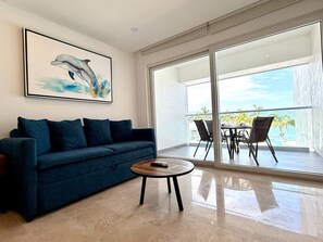 Ein modernes Wohnzimmer mit einem dunkelblauen Sofa, einem runden Couchtisch und einem großen Fenster mit Blick auf einen Balkon mit Gartenmöbeln.