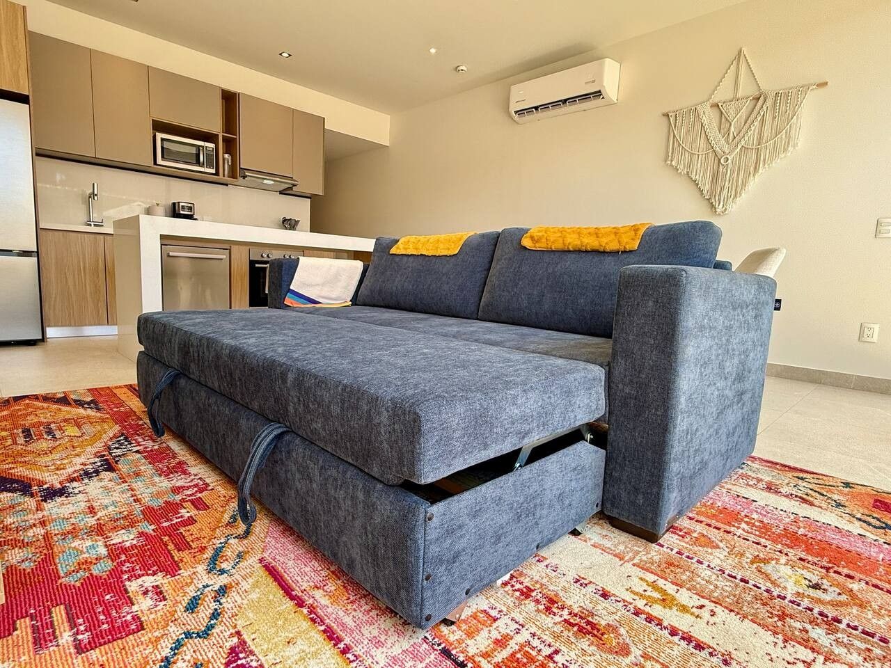 A modern living room with a grey sofa, a colorful rug, and a kitchen in the background.
