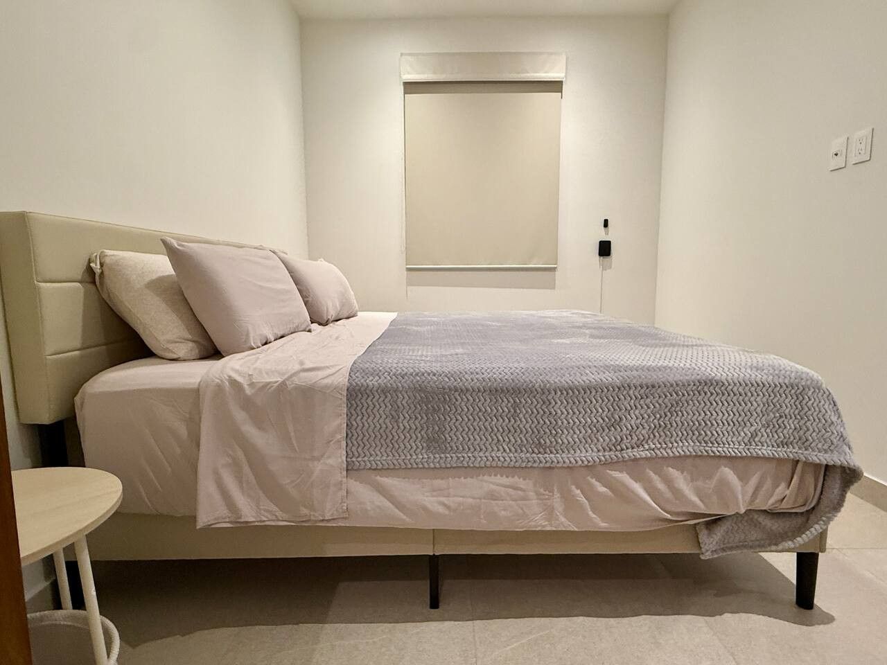 A neatly made bed with a grey blanket and white bedding, a bedside table, and a window with a blind.