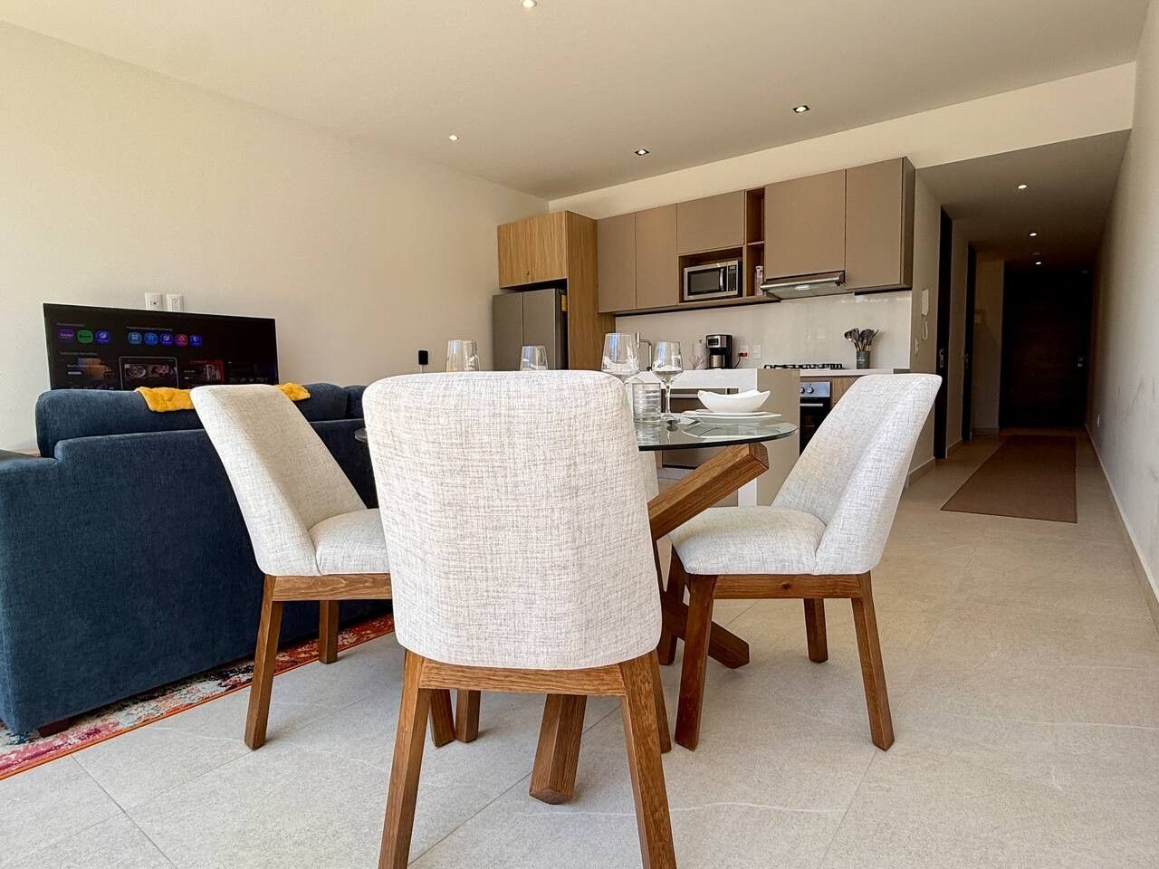 A modern living room with a sofa, a dining table with chairs, a kitchen area, and a television.