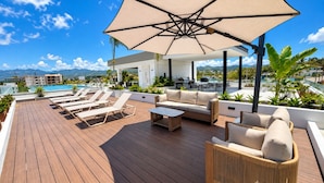 En takterrass med solstolar, en soffa och ett bord under ett stort parasoll, med utsikt över en swimmingpool och berg.