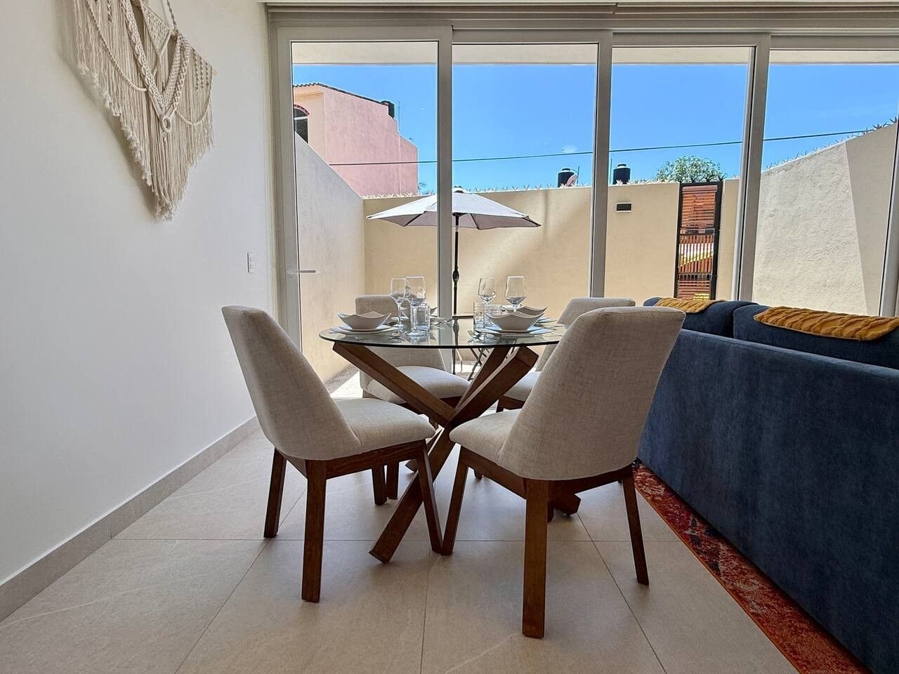 A dining area with a glass table and four chairs, a blue sofa, and a macrame wall hanging.