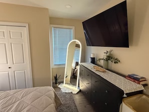 Ein Schlafzimmer mit einem Bett, einem Schminktisch, einem Spiegel, einem Fenster mit Jalousien, einem am Wand befestigten Fernseher und einer kleinen Pflanze.