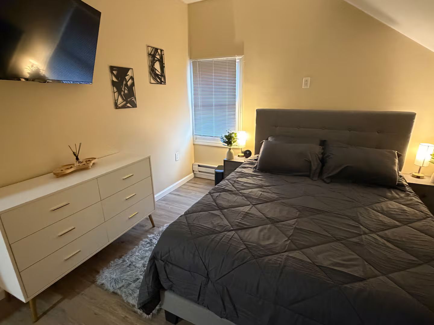 Ein Schlafzimmer mit Bett, Kommode, Nachttisch, einem an der Wand befestigten Fernseher und einem Fenster mit Jalousien.