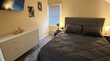 Ein Schlafzimmer mit Bett, Kommode, Nachttisch, einem an der Wand befestigten Fernseher und einem Fenster mit Jalousien.
