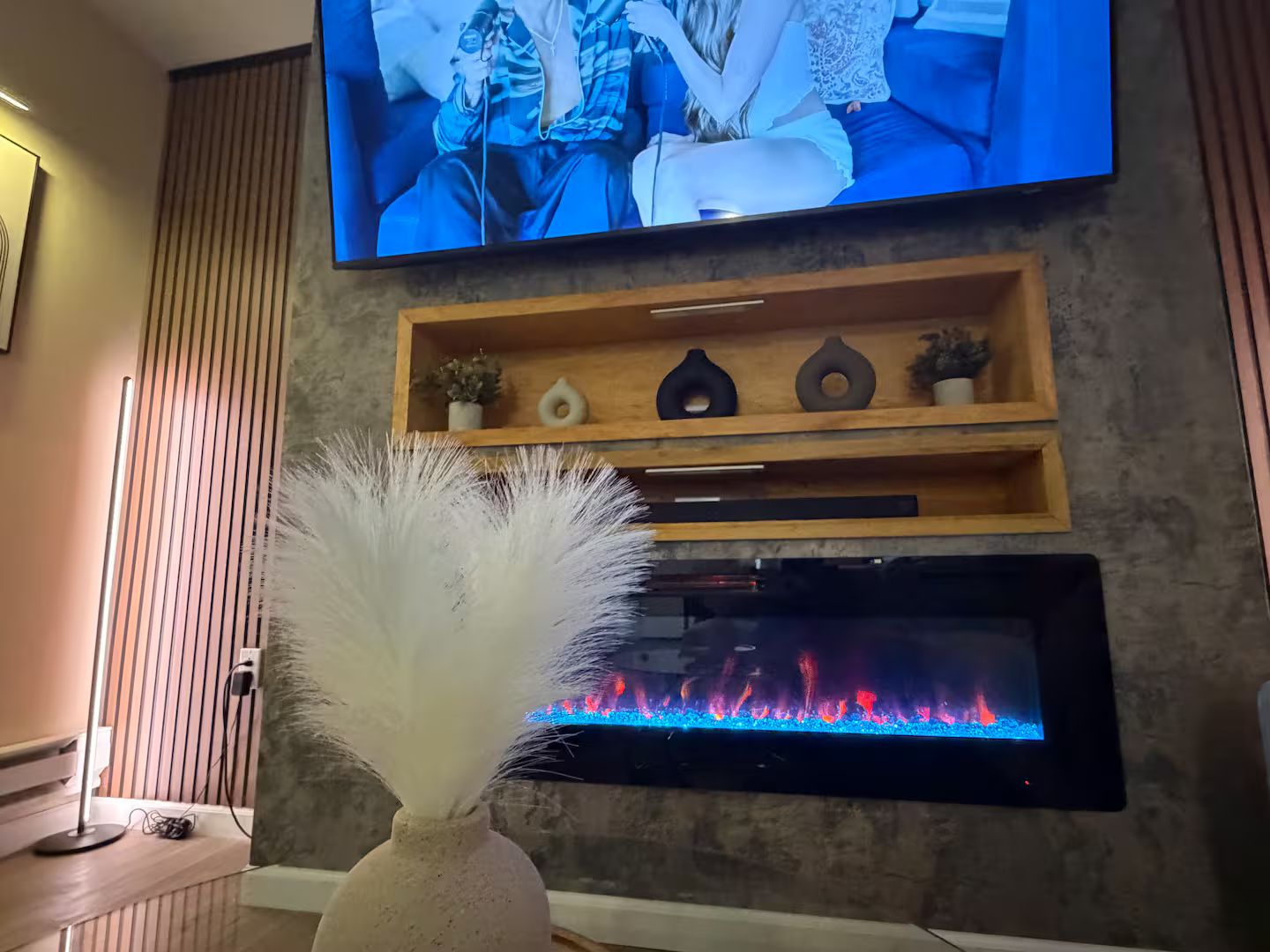 Ein Wohnzimmer mit einem an der Wand befestigten Flachbildfernseher, einem Kamin darunter und einer dekorativen Vase mit weißen Federn im Vordergrund.