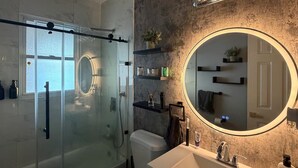 A modern bathroom with a glass-enclosed shower, a round mirror, and built-in shelves. - Spacious Luxury 3BR by Lake & Park in Roselle (Roselle)