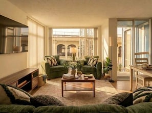 Ein Wohnzimmer mit einer Sofagarnitur, einem Couchtisch und einem Fernseher. Ein Fenster bietet Blick auf ein Gebäude.