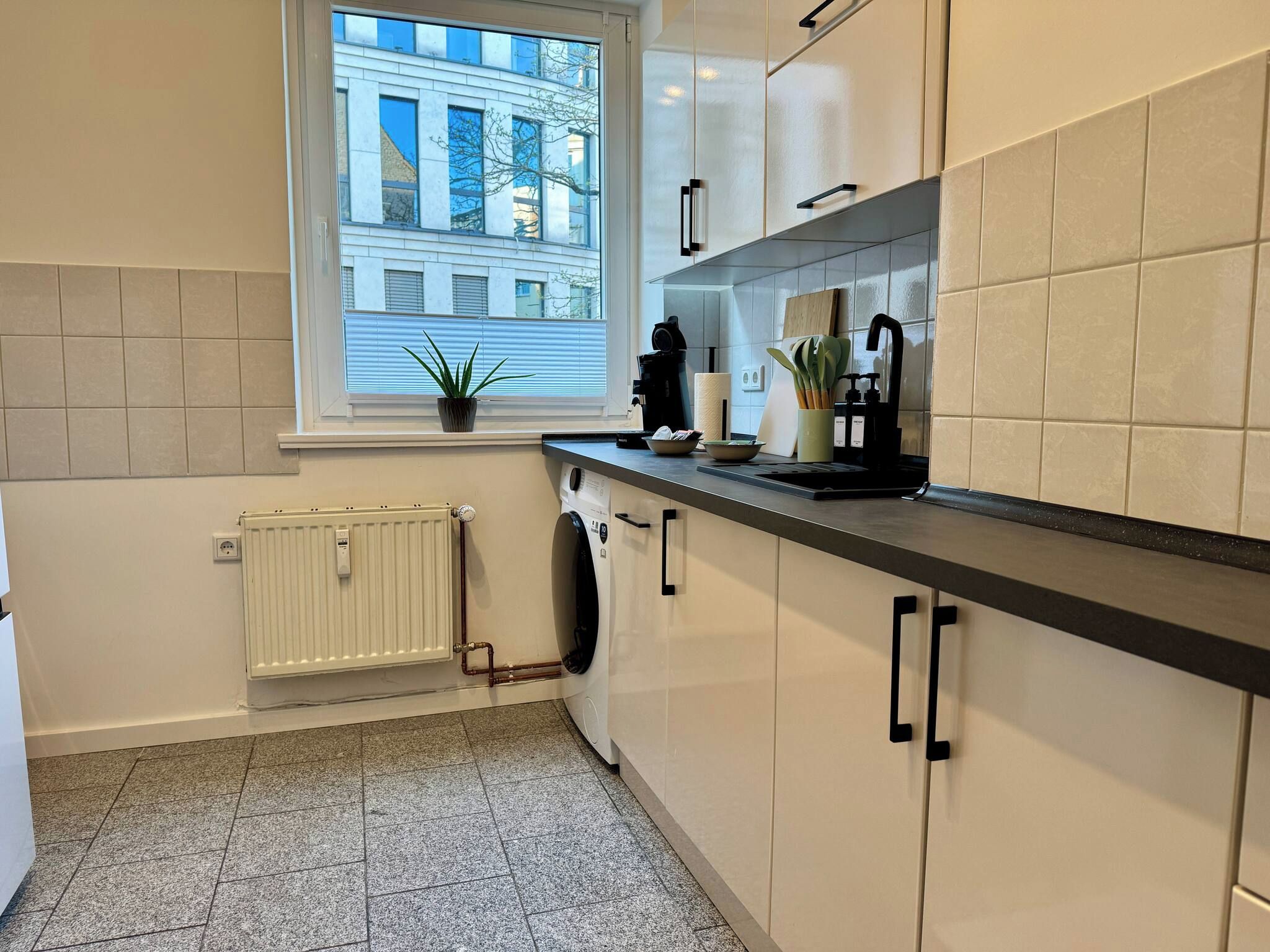 A modern kitchen with a window, a radiator, a washing machine, and a countertop with various items.