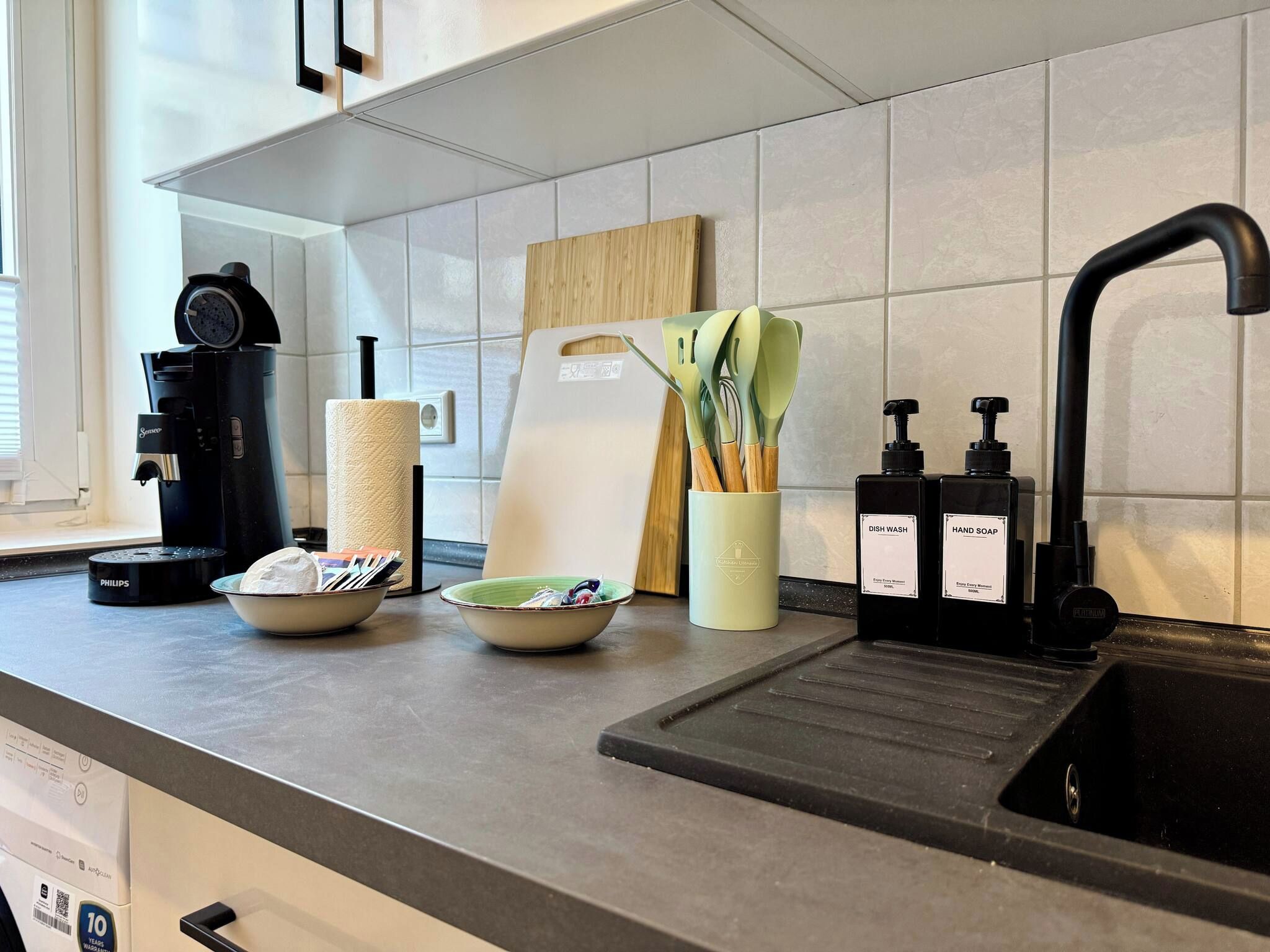 A kitchen with a coffee maker, paper towel holder, cutting board, and dish soap dispenser.